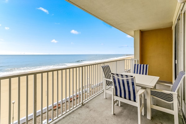 A balcony with a view of the ocean, a white table, and striped chairs.