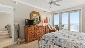 A bedroom with a large bed, a wooden dresser, a television, a ceiling fan, a large window, and a balcony with a view.