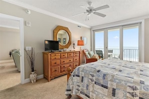 A bedroom with a large bed, a wooden dresser, a television, a ceiling fan, a large window, and a balcony with a view.