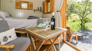 A cozy room with a bed, a wooden table with a wine bottle, and a view of greenery outside. - Moelfre - Shepherds Hut (Llanbedr)