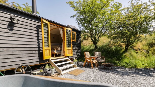 A wooden cabin with a hot tub, outdoor seating, and a gravel path leading to the entrance. - Moelfre - Shepherds Hut (Llanbedr)