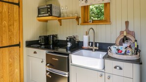 A compact kitchen with a built-in oven, a coffee maker, a sink, and a wooden shelf above the window. - Moelfre - Shepherds Hut (Llanbedr)