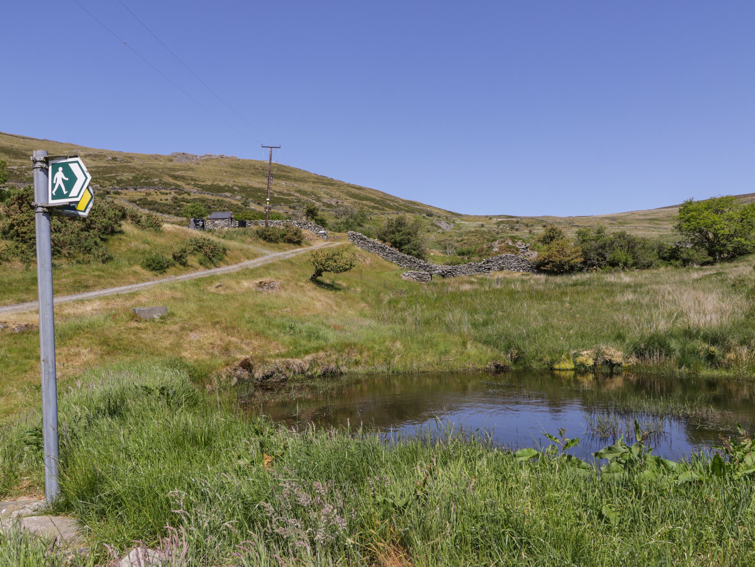 A rural landscape with a walking trail sign, a stone wall, a small pond, and a stone house in the background.