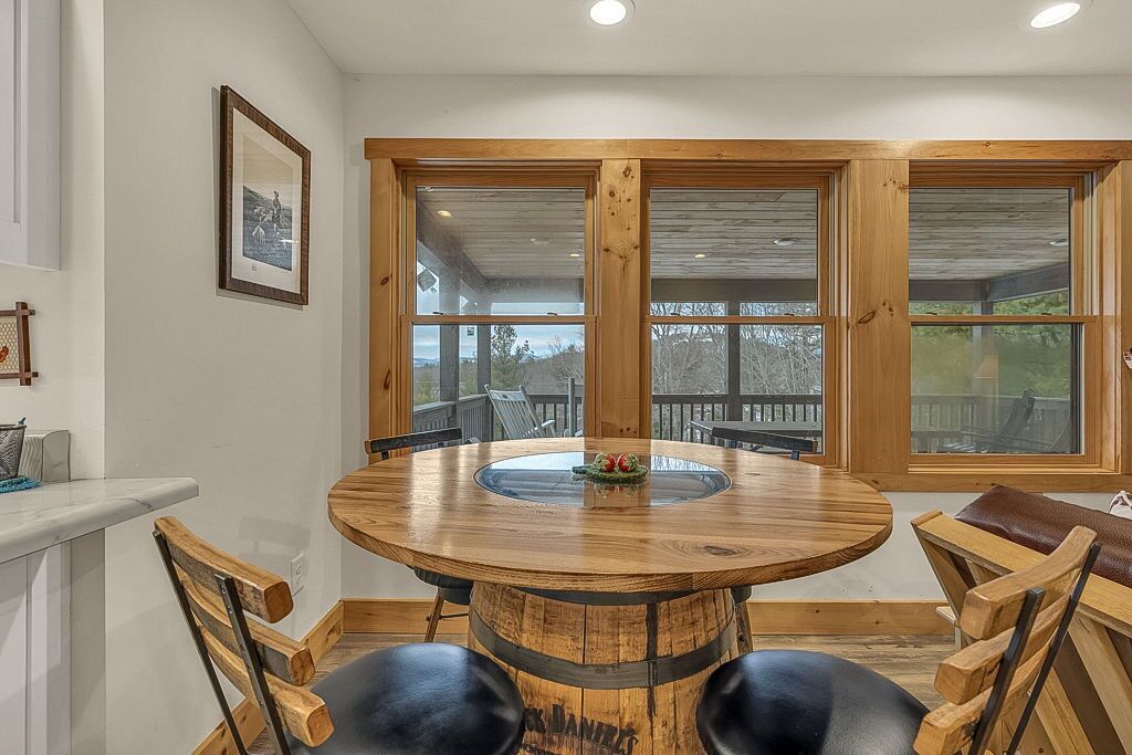 Une table ronde en bois, surmontée d’un plateau en verre, entourée de chaises en bois, dans une pièce dotée de grandes fenêtres et offrant une vue sur l’extérieur.