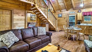 A wooden cabin interior with a brown leather sofa, a dining table with chairs, a staircase, and a kitchen area. - Fall Colors! Large Hot Tub, Private, Easy Access (Sevierville)