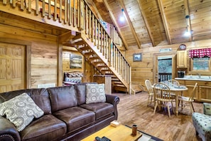 A wooden cabin interior with a brown leather sofa, a dining table with chairs, a staircase, and a kitchen area.