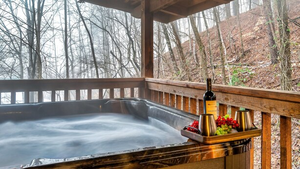A hot tub with a wooden deck and a bottle of Arrow Creek wine, surrounded by trees. - Fall Colors! Large Hot Tub, Private, Easy Access (Sevierville)