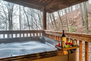 Un bain à remous avec une terrasse en bois et une bouteille de vin Arrow Creek, le tout entouré d’arbres.