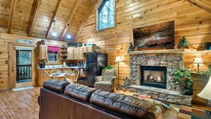 A cozy cabin interior with a stone fireplace, leather sofa, wooden dining table, and a kitchen area. - Fall Colors! Large Hot Tub, Private, Easy Access (Sevierville)