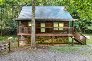 Une cabane rustique, dotée de fondations en pierre, d’une terrasse en bois et d’un toit en métal, entourée d’une forêt dense.