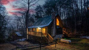 A log cabin with a wooden deck and a stone chimney, surrounded by trees and a snowy landscape. - Fall Colors! Large Hot Tub, Private, Easy Access (Sevierville)