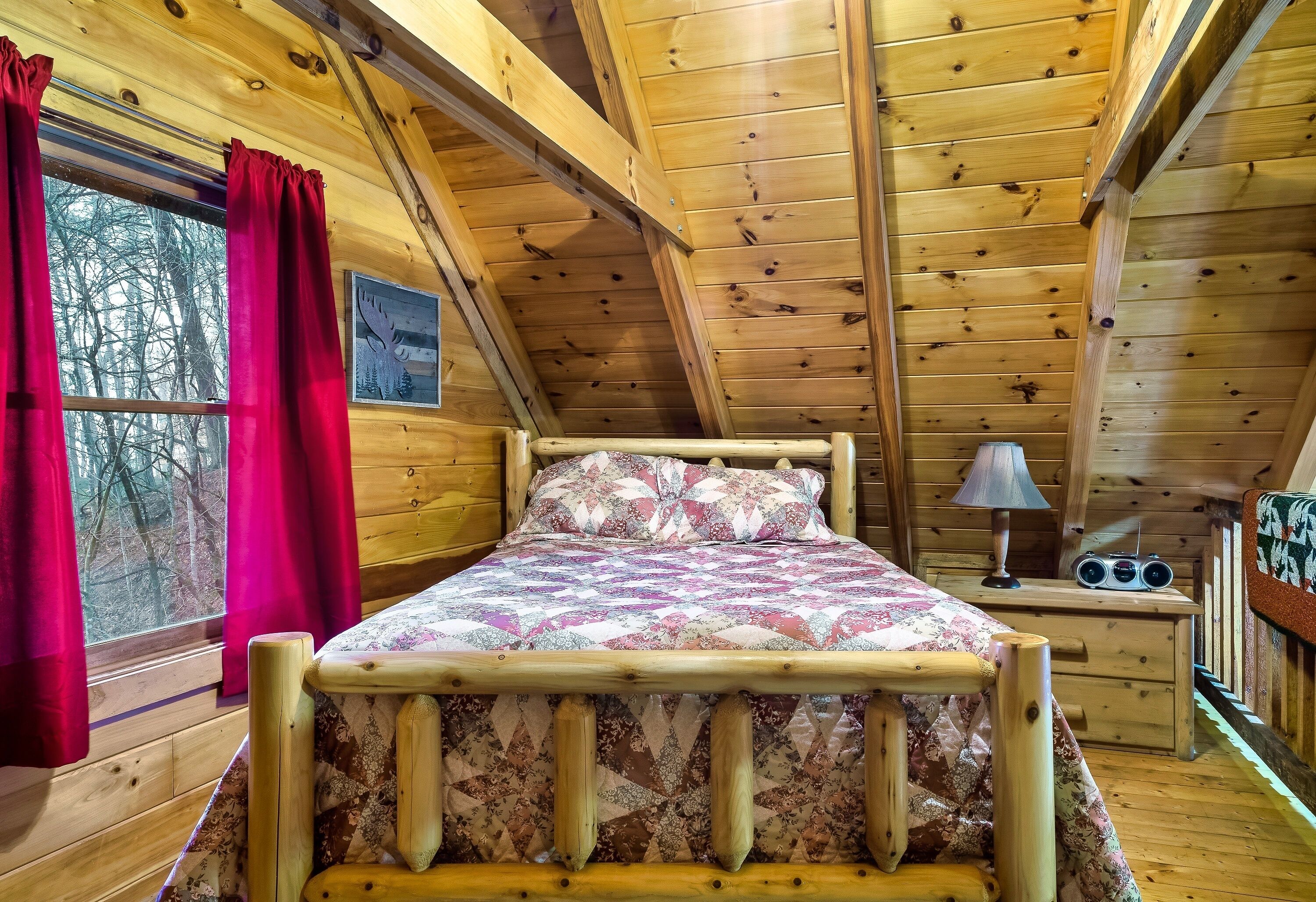 Una habitación de cabaña de madera con una cama, un ventanal con cortinas rojas, una lámpara y una radio sobre una mesita de noche.