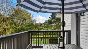 A wooden deck with a black railing, a striped umbrella, and a view of a green lawn and trees.