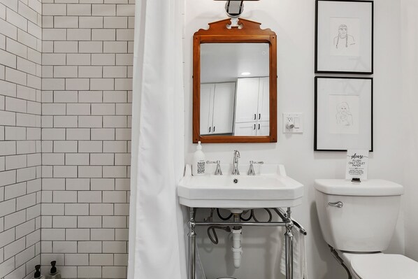 A bathroom with white tiled walls, a white sink, a wooden-framed mirror, and a toilet. There are two framed drawings on the wall.