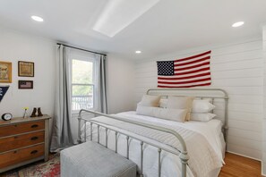 Un dormitorio con estructura de cama de metal, ropa de cama blanca, cómoda de madera, ventana con cortinas y una bandera estadounidense en la pared.