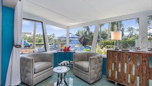 A room with two armchairs, a small round table, a wooden cabinet, and large windows offering a view of the outdoors.