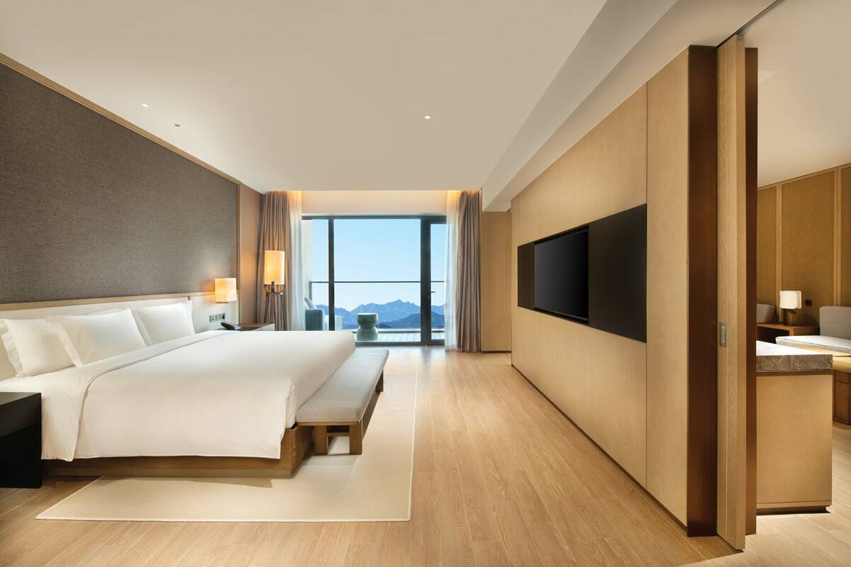 A modern hotel room with a large bed, a flat-screen TV, a wooden desk, and a view of the mountains through the window.