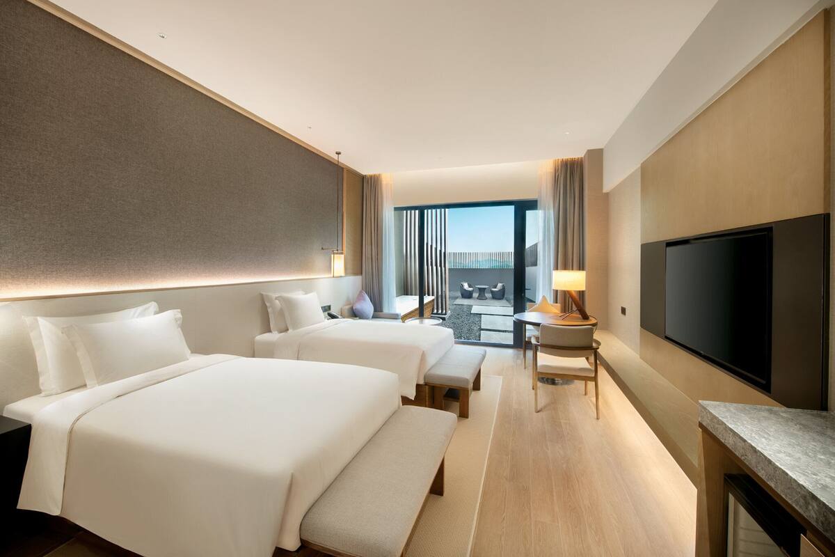 A hotel room with two beds, a TV, a desk, and a balcony with a view.