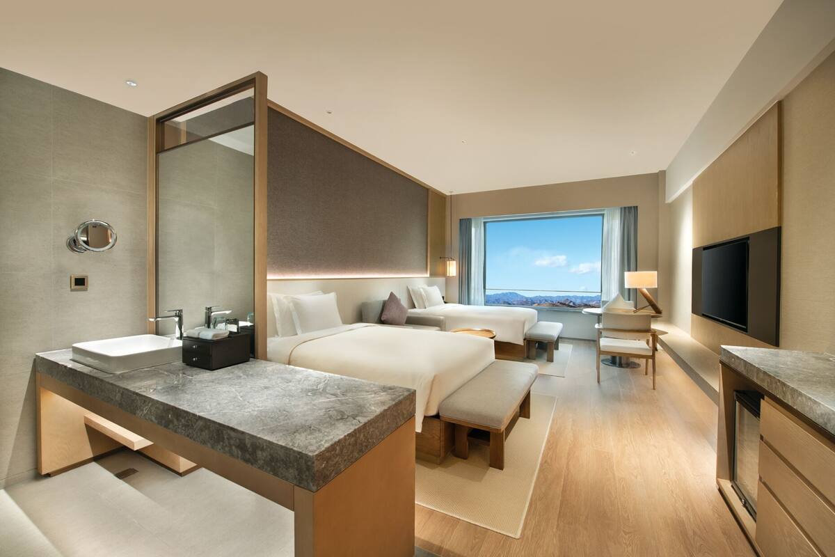 A modern hotel room with a large bed, a separate sofa, a desk with a flat-screen TV, and a bathroom with a large mirror and sink.