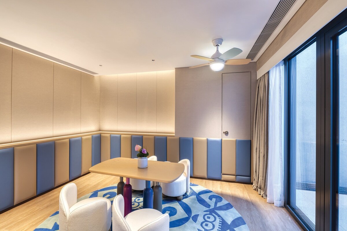 A modern hotel room with a seating area, a ceiling fan, and large windows.