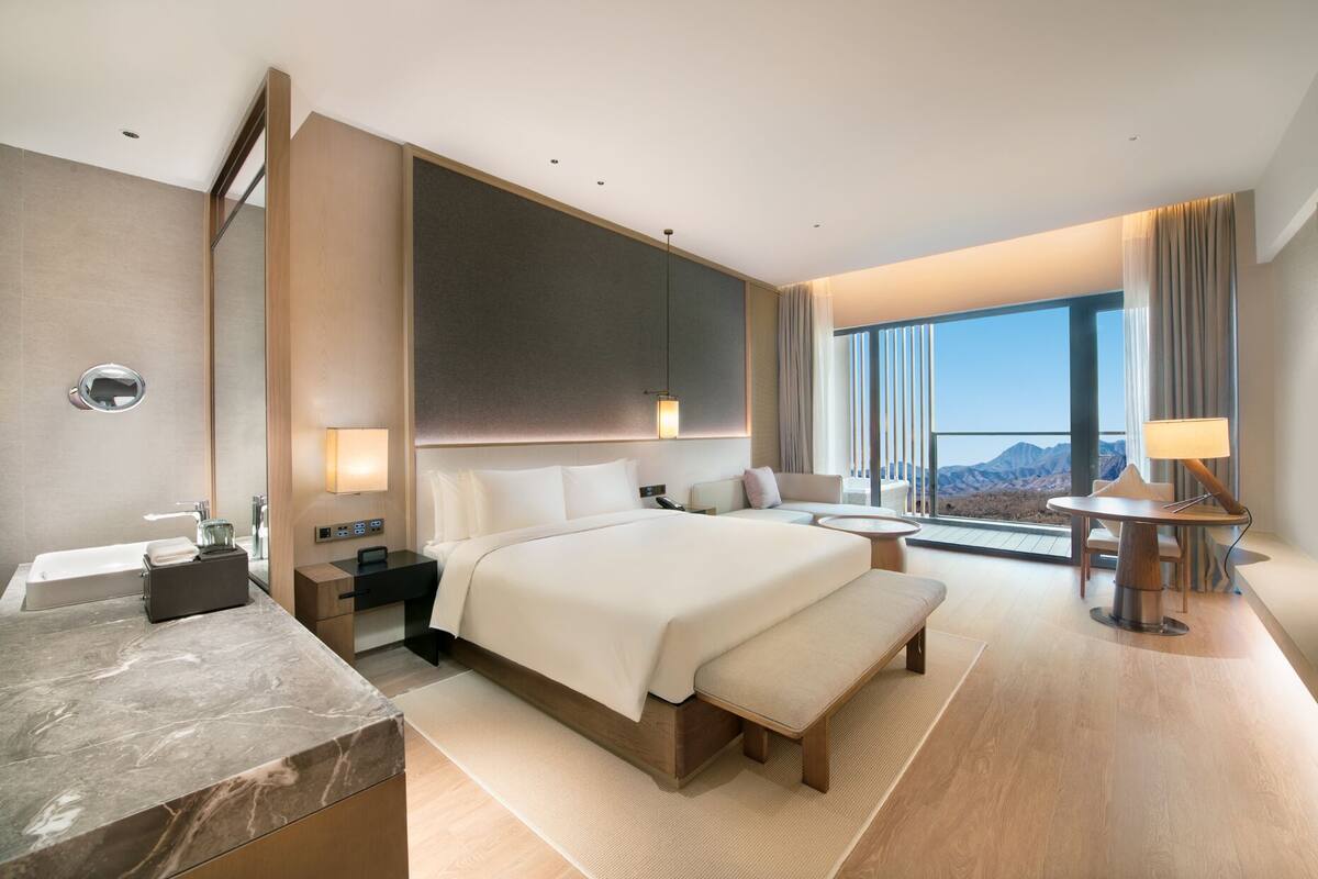 A modern hotel room with a large bed, a desk, a bathroom, and a view of mountains.