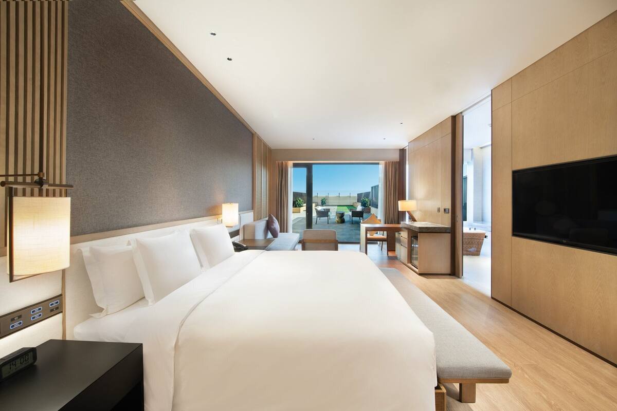 A modern hotel room with a large bed, a desk, a chair, a TV, and a view of the outdoors.