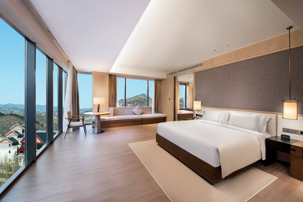 A modern hotel room with a large bed, a seating area with a view, and a desk with a lamp.