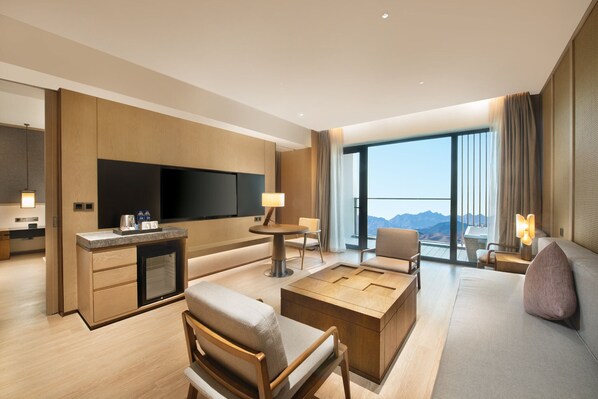 A modern hotel room with a flat-screen TV, a dining area, a sofa, and a large window with a mountain view. - Crowne Plaza Zunhua Hot Spring Town by IHG (Tangshan)