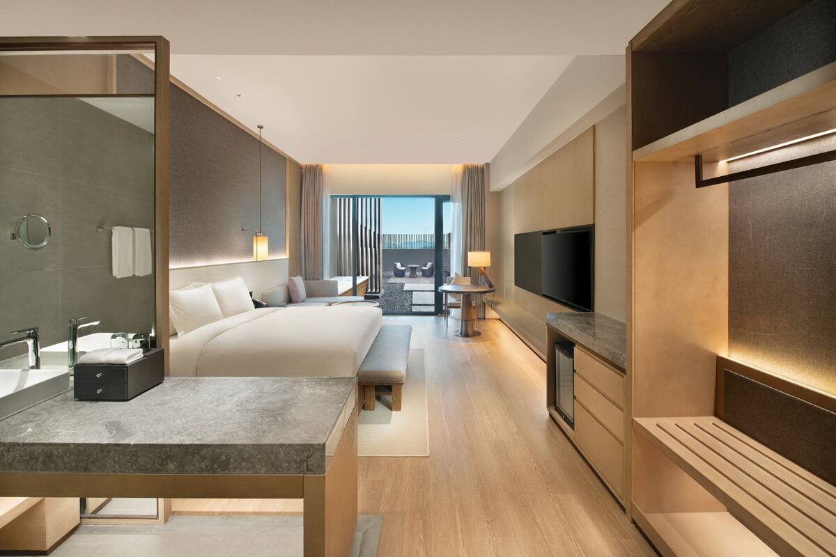 A modern hotel room with a large bed, a sitting area with a table and chairs, a TV, and a bathroom with a mirror and sink.