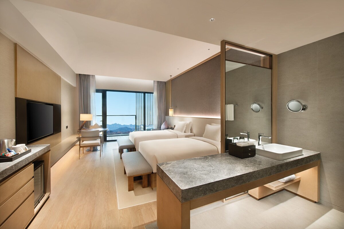 A modern hotel room with a large bed, a desk, a TV, and a bathroom with a large mirror.