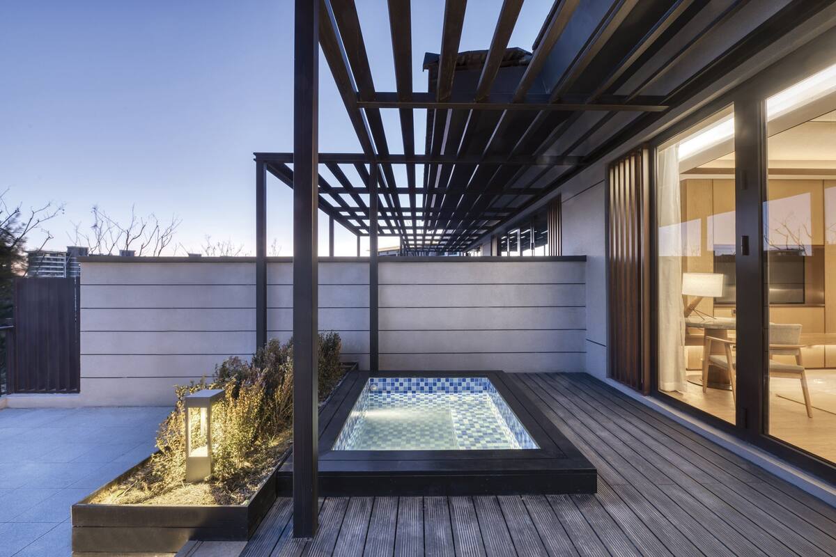 A modern outdoor hot tub area with a pergola, wooden deck, and a glass-enclosed room with furniture.