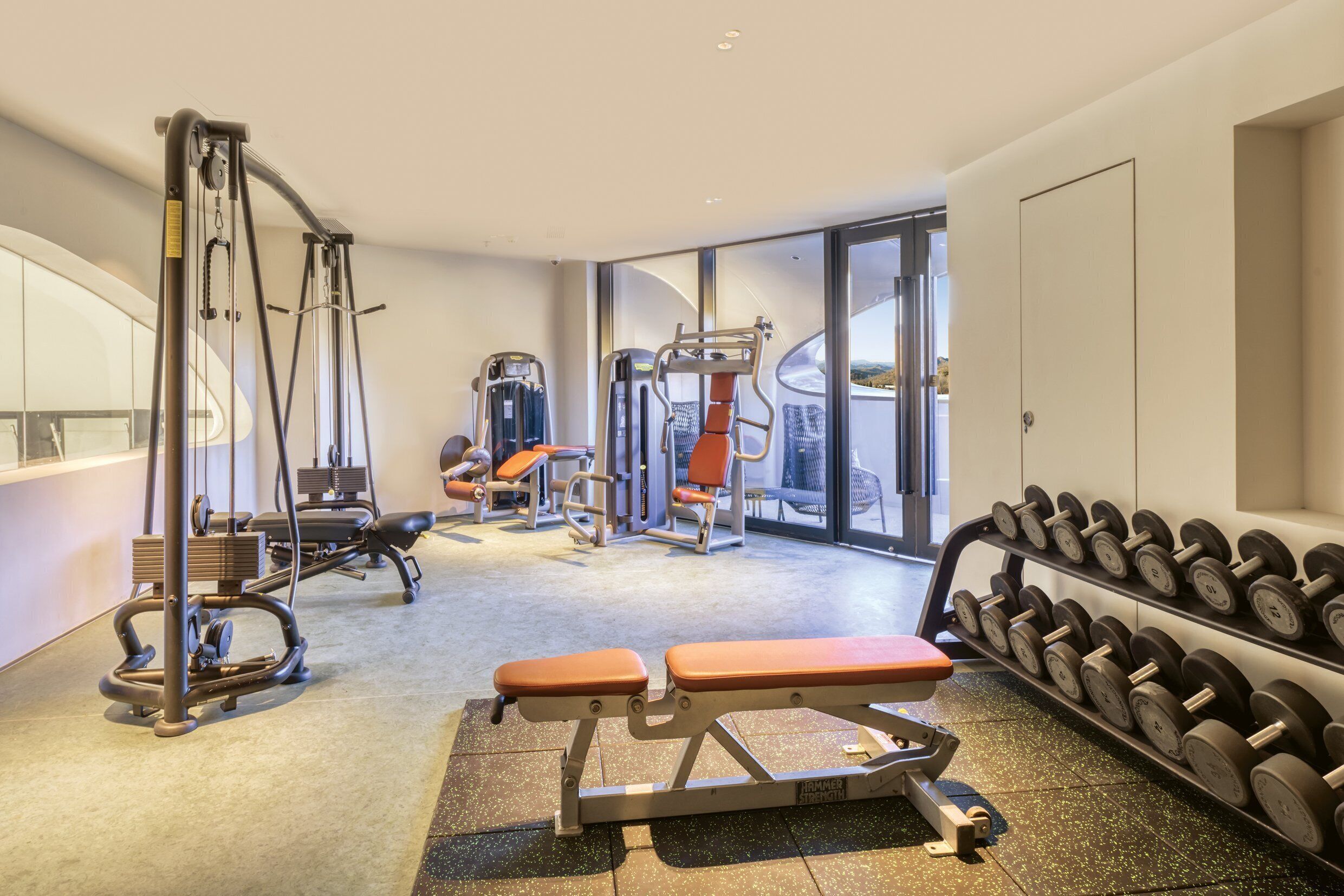 Een fitnessruimte met diverse trainingsapparaten, waaronder banken en gewichten, en een groot raam met uitzicht.