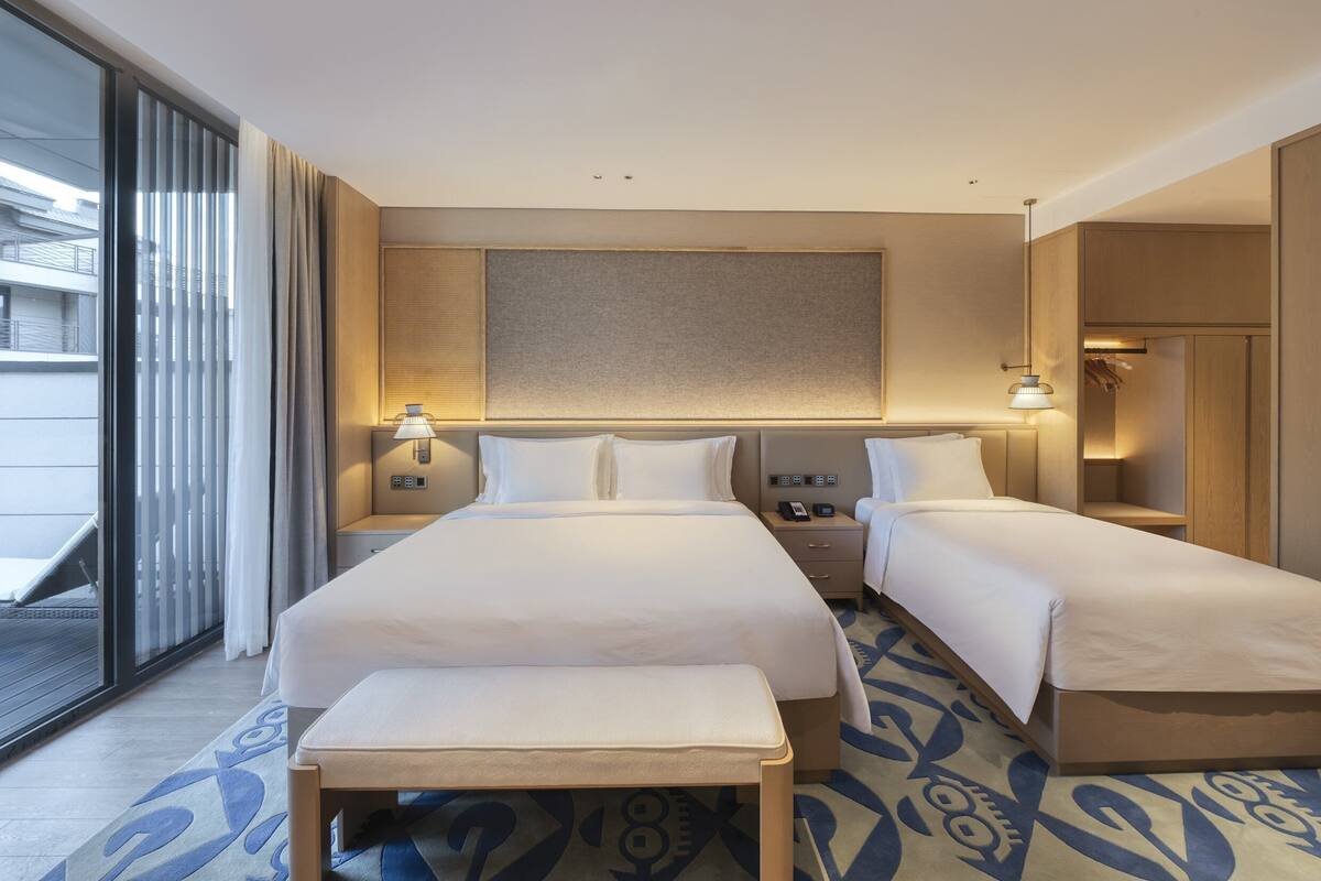 A hotel room with two beds, a bench, a large window with curtains, a bedside table with a lamp, and a wall-mounted panel.
