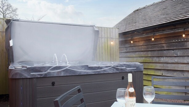 Un jacuzzi extérieur avec une terrasse en bois, une table avec une bouteille de vin et un verre, et des guirlandes lumineuses accrochées à un mur en bois.