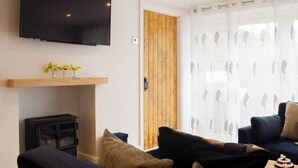 Un salon moderne avec un téléviseur à écran plat fixé au mur, une étagère en bois ornée de petites plantes, une cheminée et un coin salon confortable avec des coussins.