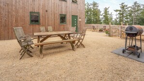 Une construction en bois avec une terrasse en gravier, une table en bois entourée de chaises et un barbecue.