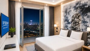 Habitación de hotel con una cama grande, vista a la ciudad a través de un ventanal y televisor de pantalla plana, además de una lámpara fijada a la pared.