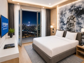 Une chambre d’hôtel avec un grand lit, une vue sur la ville par une porte vitrée, un téléviseur à écran plat et une lampe fixée au mur.