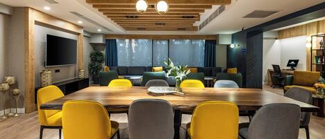 A modern conference room with a wooden table, yellow and gray chairs, a large TV, and a seating area with green and yellow sofas.