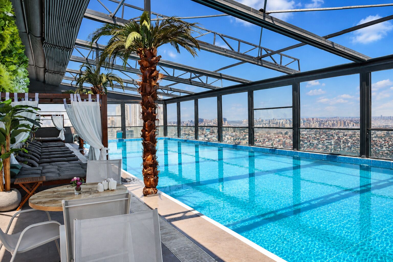 An indoor swimming pool with a city view, a palm tree, and a sunbed area.