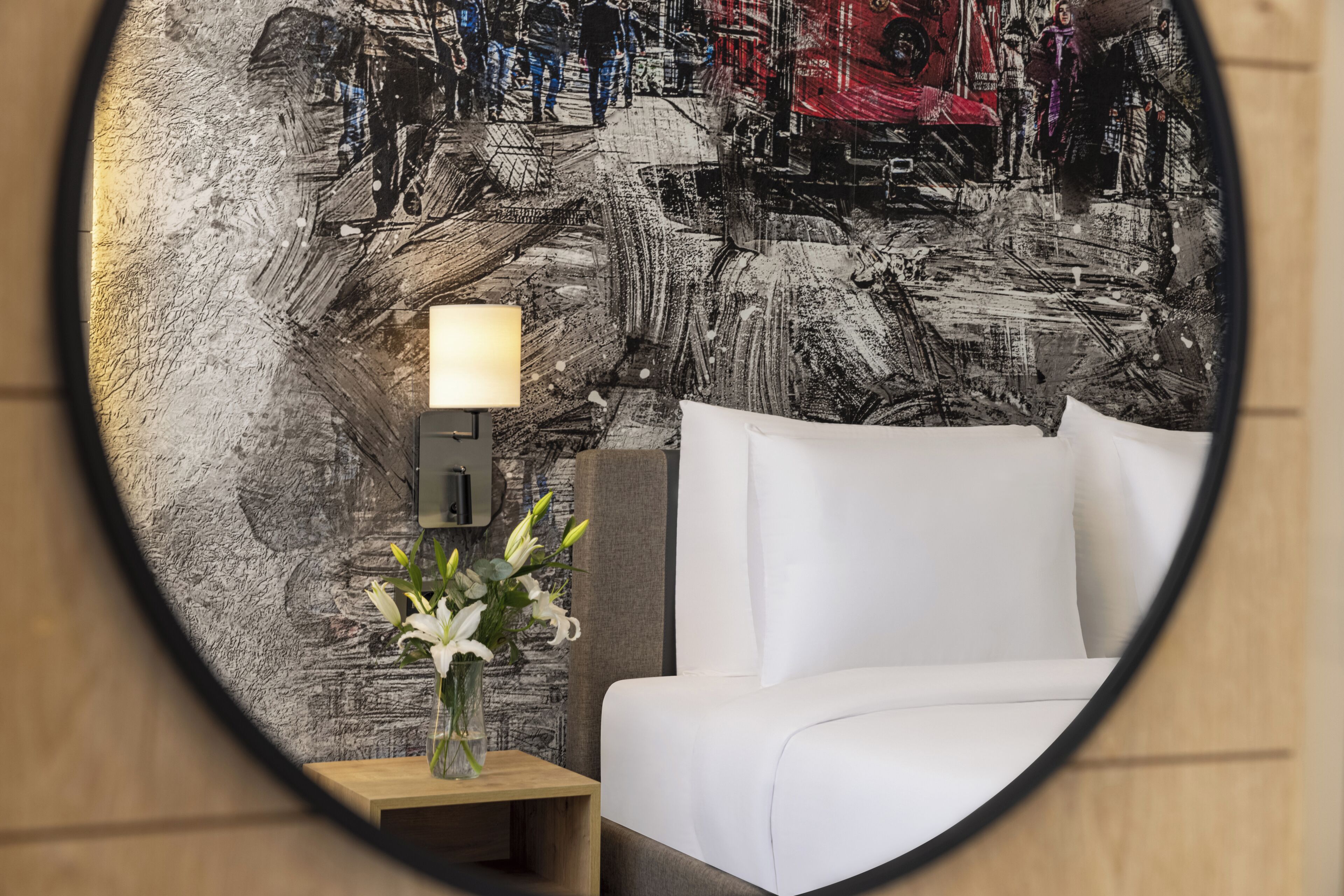 A hotel room with a bed, a nightstand with a lamp, and a wall with a textured abstract mural.