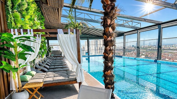 Una piscina infinita en la azotea con vista a la ciudad, un área de descanso con cojines y una mesa, y una palmera.