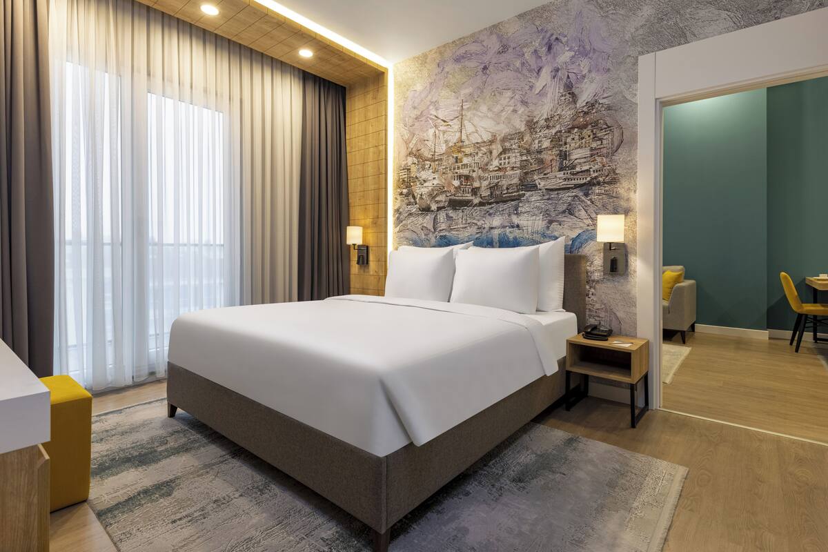 Une chambre d’hôtel moderne avec un grand lit, une fresque représentant un paysage urbain, un plafond en bois et une vue sur la ville.