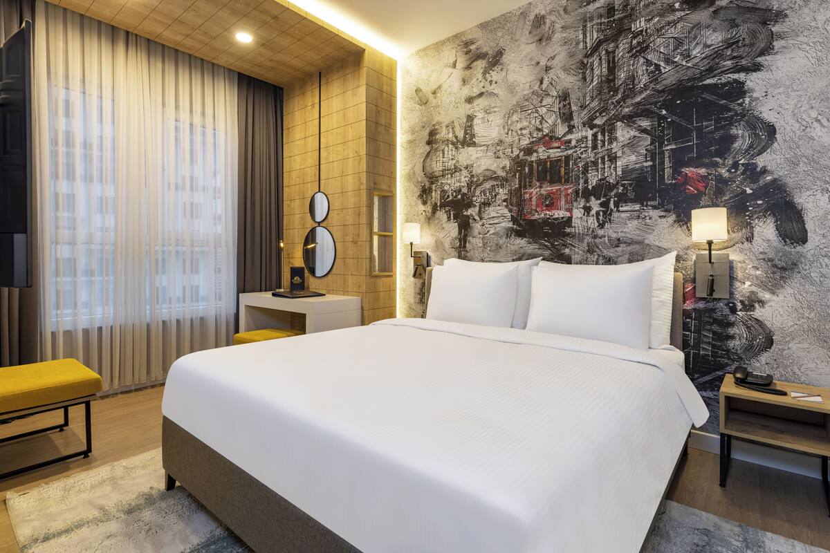 Une chambre d’hôtel moderne dotée d’un grand lit, d’un bureau, d’une chaise, d’une télévision et d’un mur orné d’une fresque représentant un paysage urbain.