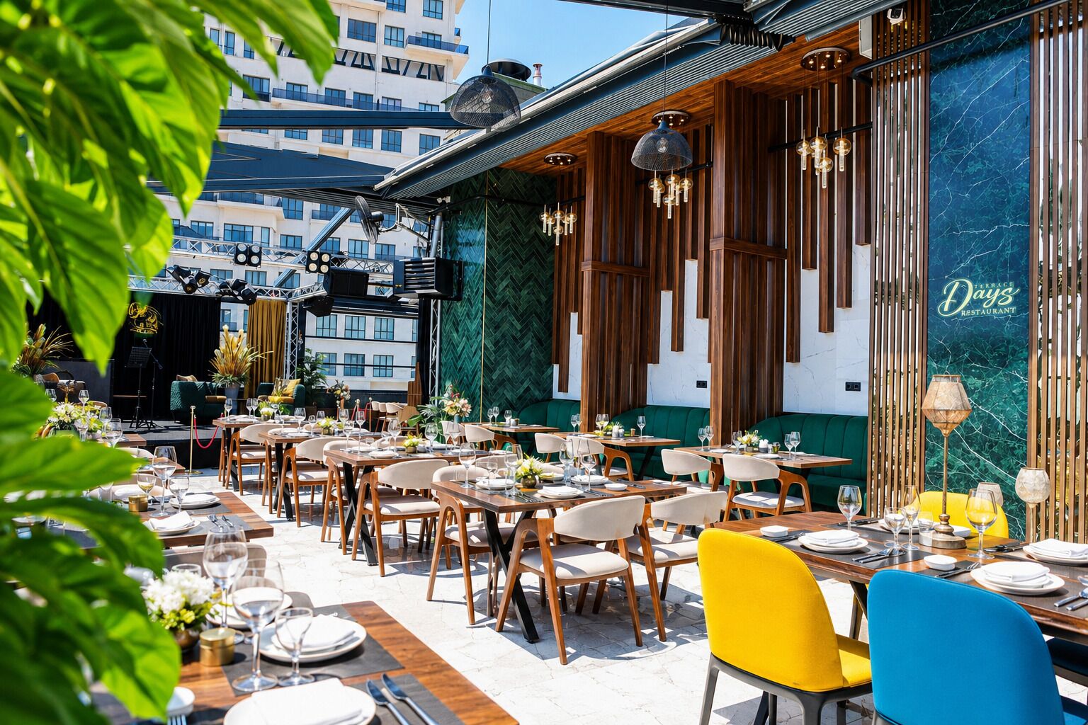Un área de comedor al aire libre con mesas y sillas, un escenario y vistas de edificios al fondo.