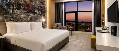 Quarto de hotel moderno com uma cama grande, uma escrivaninha, uma cadeira e vista para a cidade.