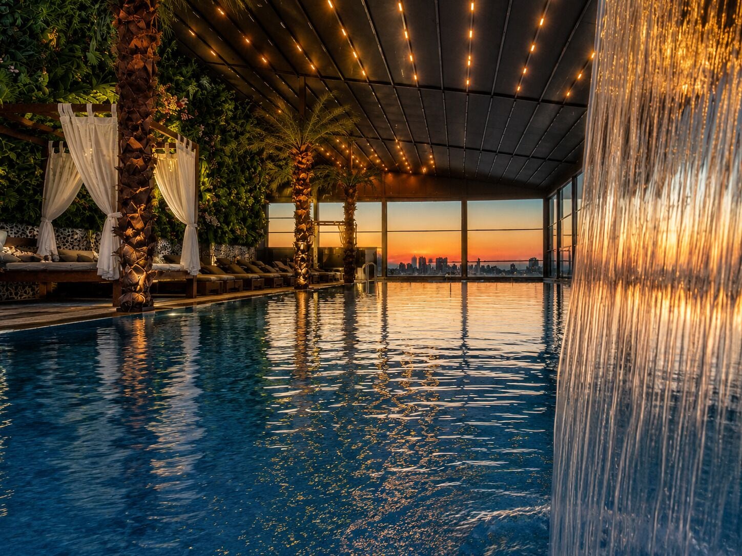An indoor swimming pool with a sunset view, palm trees, and a waterfall feature.