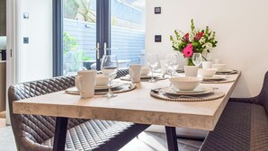 A dining table set with white plates, bowls, and glasses, a vase with flowers, and a view of the outside through a window.