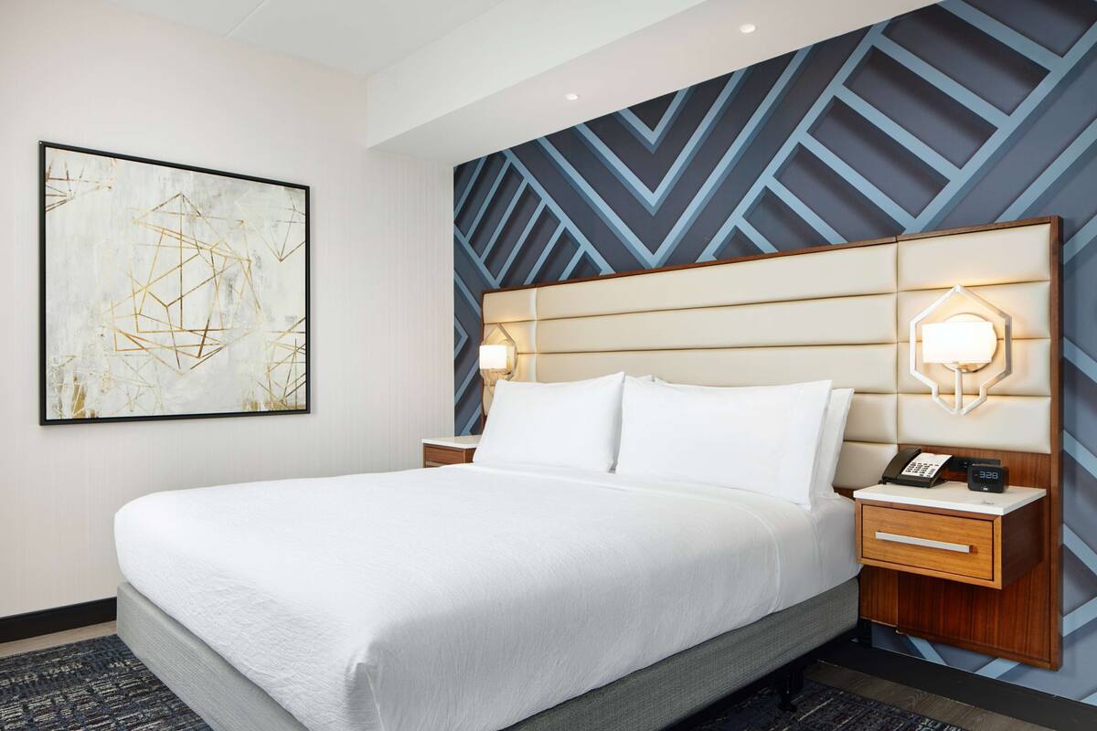 Een hotelkamer met een groot bed, twee nachtkastjes, een wandklok en een wand met een geometrisch patroon.