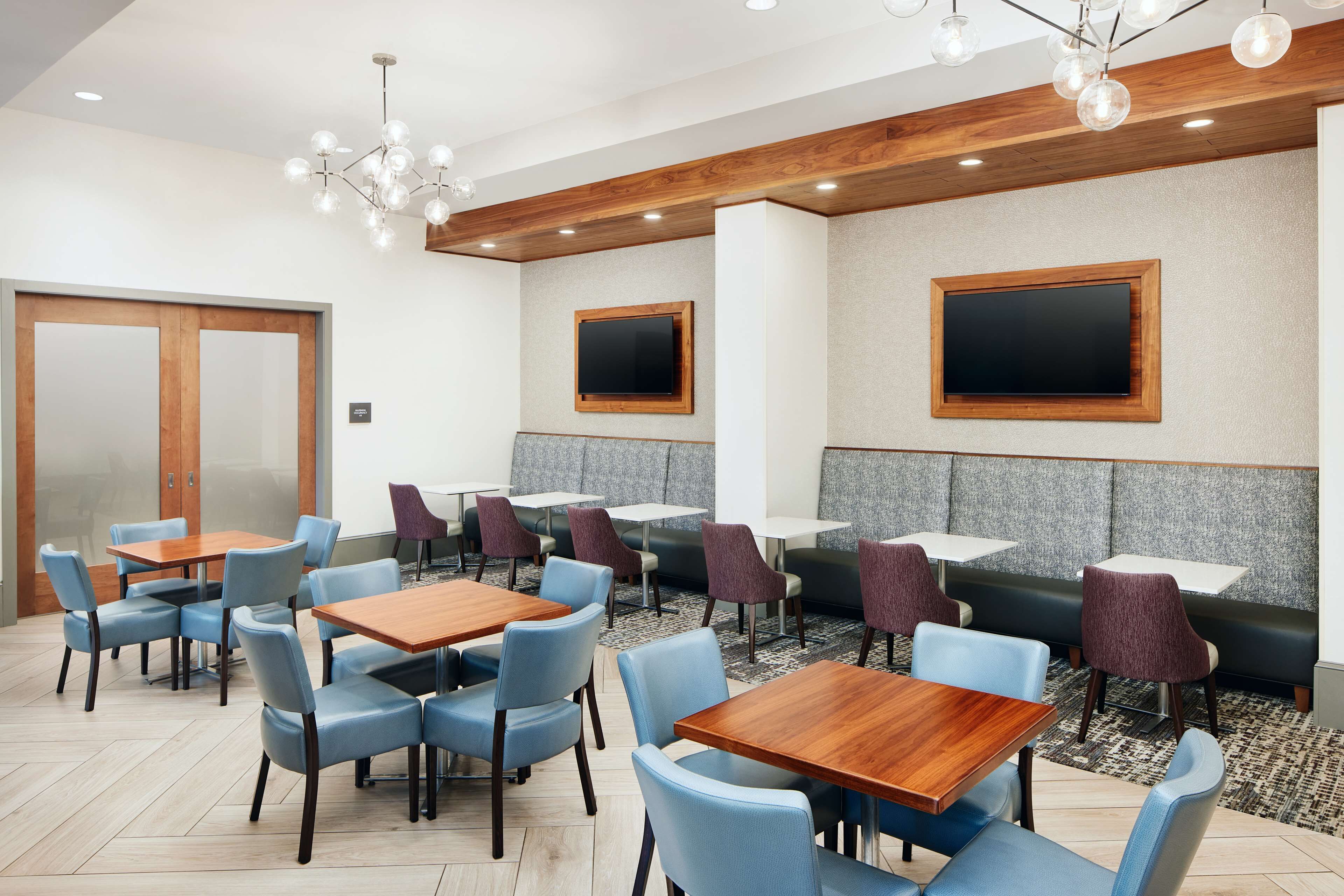 A modern restaurant with wooden tables and blue chairs, a chandelier, and two large TV screens on the walls.